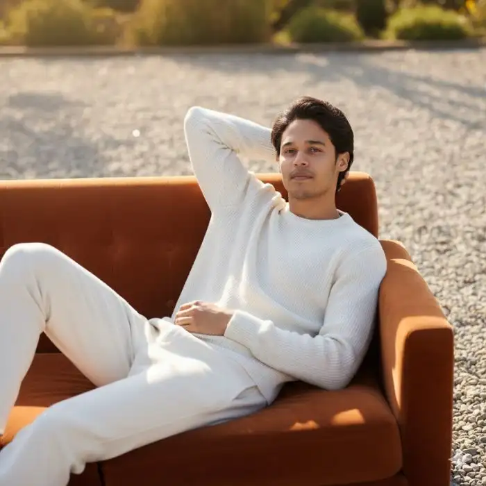 Relaxed cinematic shot of a man, Nahid Hasan Mim, wearing an off-white sweater and pants, lounging comfortably on an orange velvet sofa outdoors.